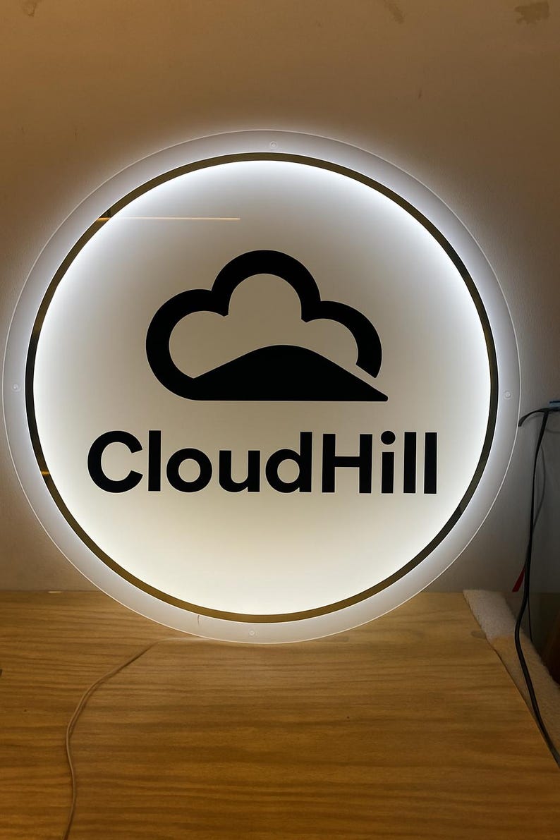 May include: Round, illuminated sign with a white background and a black cloud logo. The text "CloudHill" is displayed in black, centered below the logo. The sign has a gold-colored trim and is lit from within.
