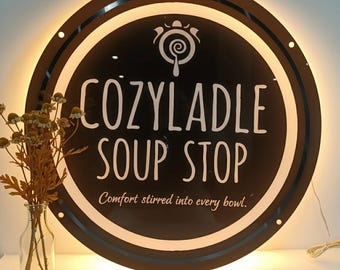May include: A circular, black, backlit sign featuring the white text COZYLADLE SOUP STOP and the slogan Comfort stirred into every bowl, with a white ladle icon above the text, positioned next to a clear glass vase with dried flowers.
