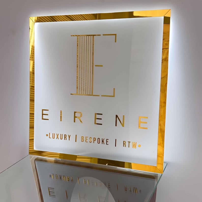 May include: A square, illuminated sign with a gold frame and a white interior. The sign features the gold text "EIRENE" and the words "LUXURY | BESPOKE | RTW." The letter "E" is stylized in gold. The sign is displayed on a glass shelf.