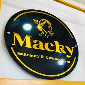 May include: A black and gold circular sign with the text "Macky - Beauty & Cosmetics" in gold lettering. A stylized image of a woman with a flower in her hair is above the text.