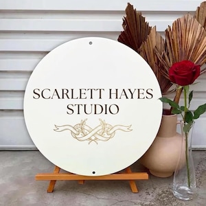 Może przedstawiać: Okrągły, kremowy znak z napisem "SCARLETT HAYES STUDIO" w kolorze brązowym. Poniżej tekstu znajduje się złoty wzór wstążki. Znak jest wyświetlany na małym drewnianym sztaludze. Czerwona róża i suszone liście są w tle.