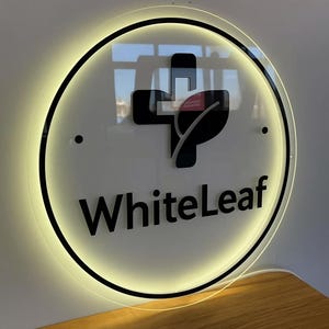 May include: Circular sign with a black border and the words "WhiteLeaf" in a bold font. The sign features a black cross and leaf design. The sign is illuminated with a warm, yellow light.