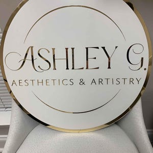 May include: A white round sign with a gold border and gold lettering. The sign reads "Ashley G. Aesthetics & Artistry".
