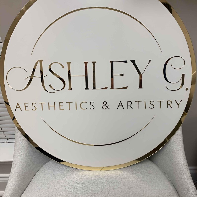 May include: A white round sign with a gold border and gold lettering. The sign reads "Ashley G. Aesthetics & Artistry".