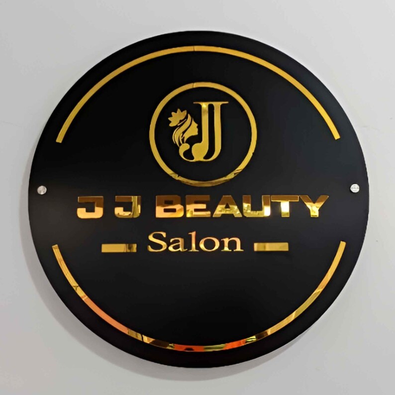 May include: A black circular sign with a gold border and gold lettering. The sign reads "JJ Beauty Salon" with a stylized gold "J" logo featuring a woman's silhouette with a crown.
