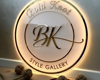 Puede incluir: Un letrero redondo e iluminado con el texto "Bold Knot Style Gallery" en letras doradas. El letrero presenta las letras "BK" en una fuente estilizada. Nudos de cuerda decorativos y lazos de cuero en primer plano.