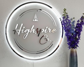 Puede incluir: Un letrero redondo e iluminado con el texto "Highspire Co." en una fuente elegante y cursiva. El letrero es plateado y tiene una luz blanca brillante alrededor del borde. Un jarrón de flores moradas y azules está a la derecha.
