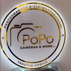 May include: Circular sign with a gold border and the words "PHOTOGRAPHER & CINEMATOGRAPHERS" around the top. The center features a camera graphic and the word "PoPo" in gold, with "CAMERAS & MORE..." below. "TRUSTED CHOICE" is at the bottom.