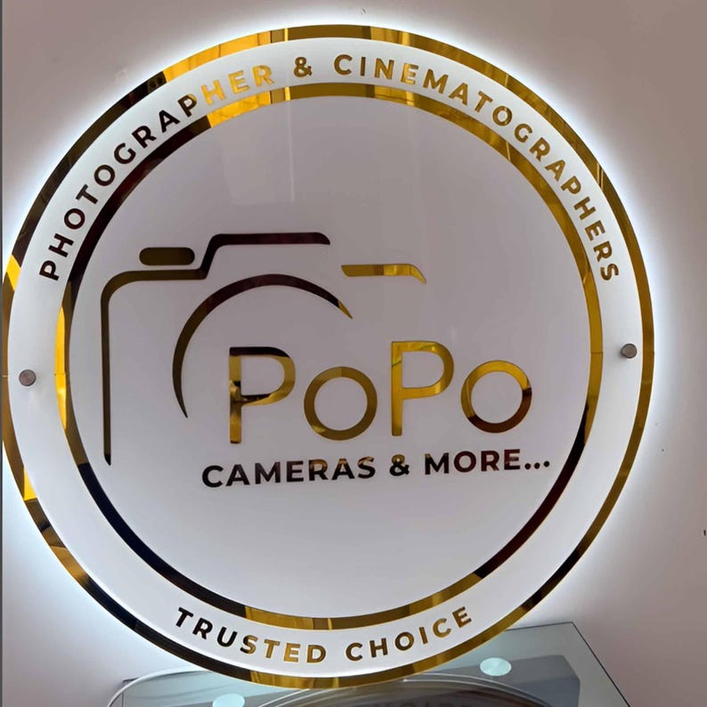 May include: Circular sign with a gold border and the words "PHOTOGRAPHER & CINEMATOGRAPHERS" around the top. The center features a camera graphic and the word "PoPo" in gold, with "CAMERAS & MORE..." below. "TRUSTED CHOICE" is at the bottom.