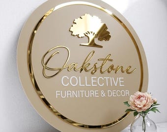 Puede incluir: Un letrero redondo con borde dorado y el texto "Oakstone Collective Furniture & Decor". El letrero presenta un emblema de árbol dorado y letras doradas en cursiva. Un pequeño jarrón con una flor rosa está en primer plano.
