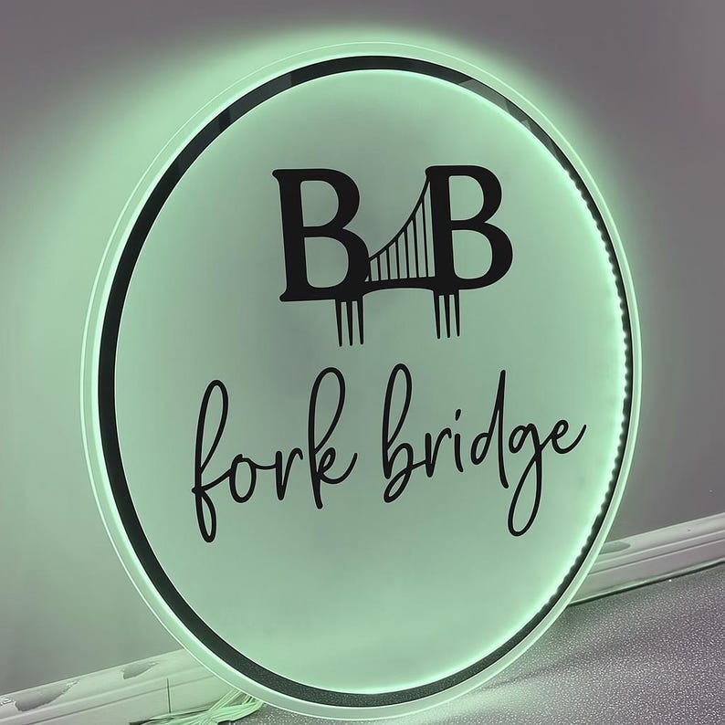 May include: Round, illuminated sign with a black border and a light green glow. The sign features the text "B B" above a bridge graphic, and the words "fork bridge" in a cursive font. The sign is likely for a business.