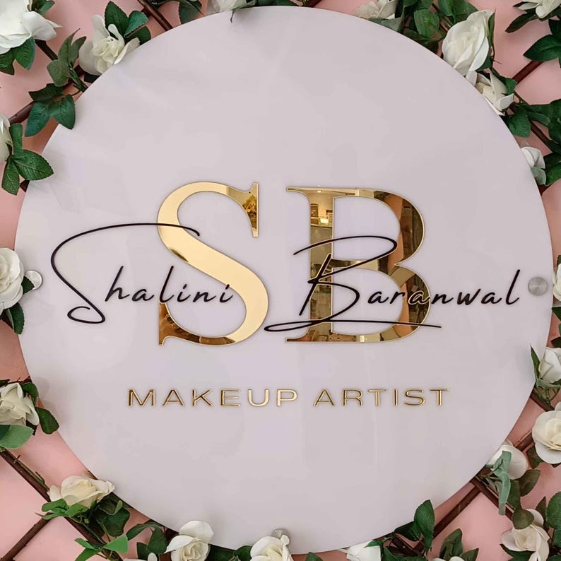 May include: A white round sign with gold lettering that reads "SB Shalini Baranwal MAKEUP ARTIST". The sign is decorated with white flowers.