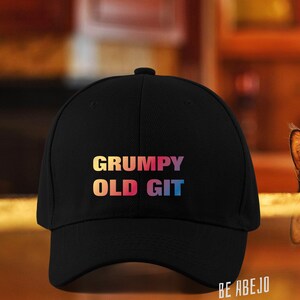 Può includere: Un cappello da baseball nero con il testo "GRUMPY OLD GIT" in colori arcobaleno. Il cappello è su un tavolo con un gatto nero che sporge dal bordo.