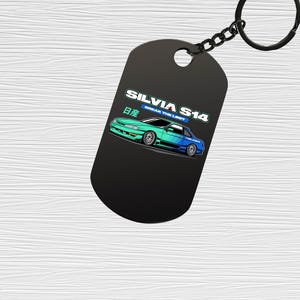 Puede incluir: Llavero con etiqueta negra con un diseño gráfico de un coche turquesa y azul. La etiqueta tiene el texto "SILVIA S14" y "BREAK THE LIMIT". El llavero tiene un anillo y una cadena negros.