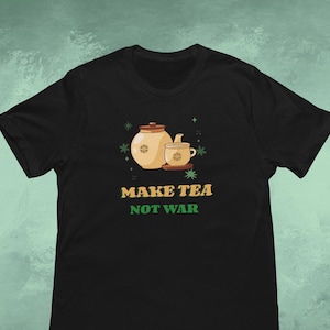 Pode incluir: Camiseta preta com um gráfico de conjunto de chá, incluindo uma chaleira e uma xícara. O texto "MAKE TEA NOT WAR" é impresso em verde e dourado. O design inclui estrelas, criando um tema pacífico.