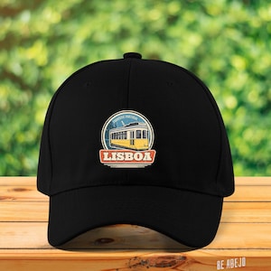 Op de afbeelding: Zwarte baseballpet met een geborduurde patch in vintage stijl met een gele tram en de tekst "Touristic Portugal Lisboa".