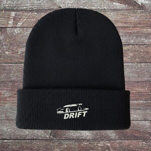 May include: Black beanie with a white graphic of a car and the word "DRIFT" in white. The beanie is made of a knitted material and has a folded brim. The design is centered on the front of the hat.
