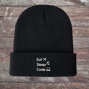 Puede incluir: Gorro de punto negro con la inscripción blanca "Eat Sleep Code", acompañada de un tenedor y un cuchillo, un rayo y un icono de portátil. El borde del gorro está doblado.