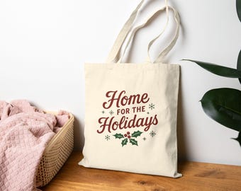 May include: Cream-colored tote bag with the text "Home for the Holidays" in burgundy and green. The design includes snowflakes and holly leaves. The bag has two handles and is made of a canvas-like material.