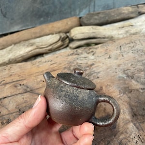 Könnte beinhalten: Kleine, braune Keramik-Teekanne mit passendem Deckel. Die Teekanne hat einen runden Körper, einen gebogenen Henkel und einen Ausguss. Die Oberfläche hat ein strukturiertes, gealtertes Aussehen. Die Teekanne wird in einer Hand vor einem Hintergrund aus Treibholz gehalten.