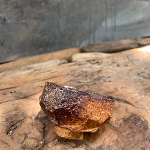 May include: A small, handmade ceramic bowl with a rough, textured exterior. The bowl is primarily brown with areas of darker, almost black, coloration. It sits on a small, circular base, also brown. The bowl is displayed on a weathered wooden surface.