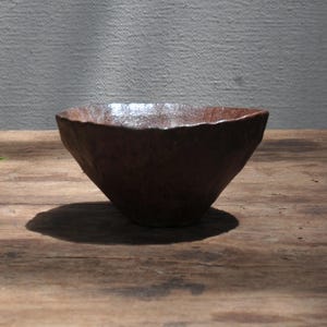 May include: A small, dark brown ceramic bowl with a textured, uneven surface and a wide, flared rim. The bowl features a deep, conical shape with a rough, hand-crafted appearance and a subtle sheen on the interior rim, reflecting light.