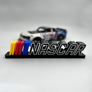 Puede incluir: Un logotipo NASCAR impreso en 3D en negro con una base negra y rayas de colores. El logotipo está sobre una superficie blanca.