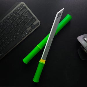 Puede incluir: Una espada de juguete verde y plateada con un mango de bambú. La espada está sobre una superficie negra con un teclado y un ratón en el fondo.