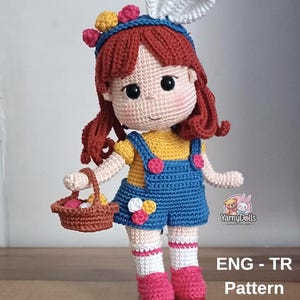 May include: A handmade crochet doll with red hair, wearing a blue headband with bunny ears and a blue denim overall dress. The doll holds a basket with colorful eggs. The doll's outfit includes a yellow shirt, pink shoes, and white socks.