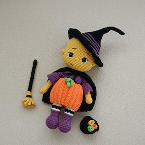 May include: A crocheted doll with yellow skin, wearing a black and purple pointed hat, a purple shirt, and an orange pumpkin-shaped skirt with a black cape. The doll is accompanied by a small black broom with yellow bristles and a black cauldron.