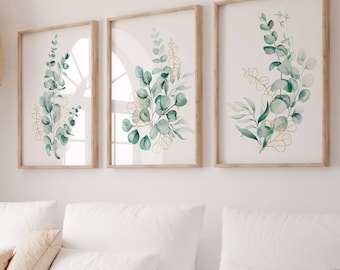 Puede incluir: Un sofá seccional blanco con almohadas y una manta beige, una pantalla de lámpara tejida colgando arriba y tres impresiones de acuarela enmarcadas de hojas de eucalipto verdes en una pared blanca.