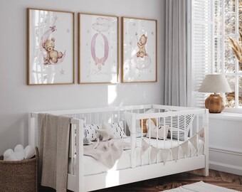 May include: Three watercolour prints featuring a pink cloud with a teddy bear holding a star, a pink letter O with a bow, and a pink moon with a teddy bear sleeping. The prints are framed in wood and hung on a white wall above a white cot.