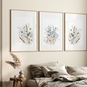 May include: Three framed prints of watercolour paintings of blue and white wildflowers with brown stems. The prints are arranged in a row on a light beige wall.