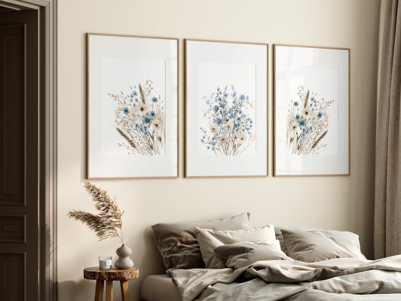 May include: Three framed prints of watercolour paintings of blue and white wildflowers with brown stems. The prints are arranged in a row on a light beige wall.