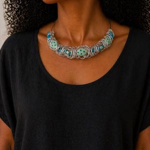 May include: A silver wire necklace featuring blue and green beads. The necklace has a distinctive design, with the beads enclosed in a woven wire structure. The beads are a mix of solid blue and green with a star pattern. The necklace is worn against a black top.