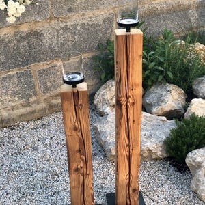K&ouml;nnte beinhalten: Zwei rustikale Kerzenhalter. Jeder hat einen transparenten Glaszylinder auf einem schwarzen Metallsockel, der auf einem verwitterten, vertikalen Holzpfosten montiert ist. Das Holz zeigt Maserung und Knoten. Die Kerzenhalter stehen auf einem Bett aus kleinen Steinen, mit einer Steinmauer im Hintergrund.