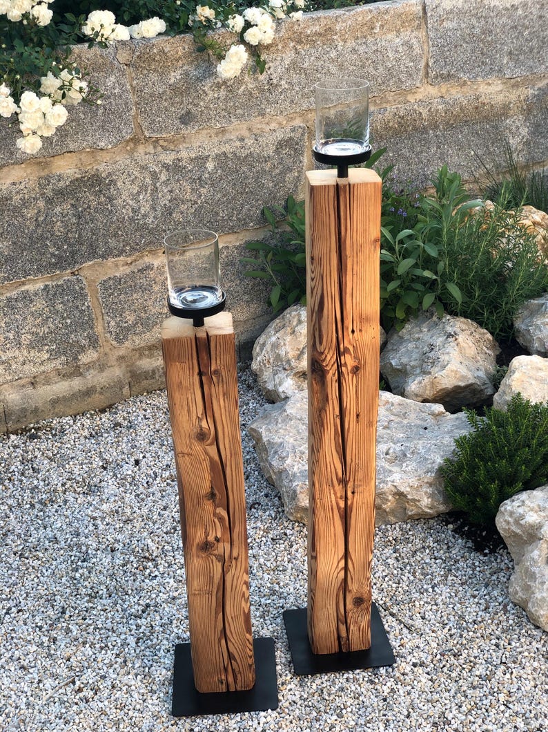 K&ouml;nnte beinhalten: Zwei rustikale Kerzenhalter. Jeder hat einen transparenten Glaszylinder auf einem schwarzen Metallsockel, der auf einem verwitterten, vertikalen Holzpfosten montiert ist. Das Holz zeigt Maserung und Knoten. Die Kerzenhalter stehen auf einem Bett aus kleinen Steinen, mit einer Steinmauer im Hintergrund.