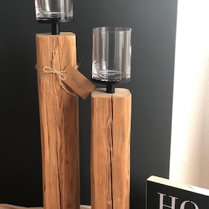 K&ouml;nnte beinhalten: Zwei Holzkerzenhalter mit klaren Gl&auml;sern. Die Halter sind aus hellbraunem Holz und haben einen schwarzen Metallsockel. Ein kleines Etikett mit dem Wort "Herzen" ist an einem der Halter befestigt. Ein schwarz-wei&szlig;es Schild mit der Aufschrift "HOME" befindet sich im Hintergrund.