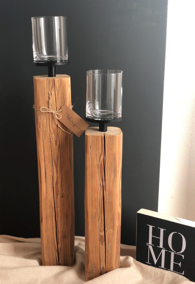 K&ouml;nnte beinhalten: Zwei Holzkerzenhalter mit klaren Gl&auml;sern. Die Halter sind aus hellbraunem Holz und haben einen schwarzen Metallsockel. Ein kleines Etikett mit dem Wort "Herzen" ist an einem der Halter befestigt. Ein schwarz-wei&szlig;es Schild mit der Aufschrift "HOME" befindet sich im Hintergrund.
