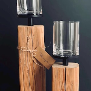 K&ouml;nnte beinhalten: Zwei Kerzenhalter aus Holz mit transparenten Glas-Windlichtern. Die Holzsockel sind rechteckig und haben eine nat&uuml;rliche Holzmaserung. Die Schirme sitzen auf schwarzen Metallplattformen. Ein braunes Etikett mit Text ist an einem befestigt.