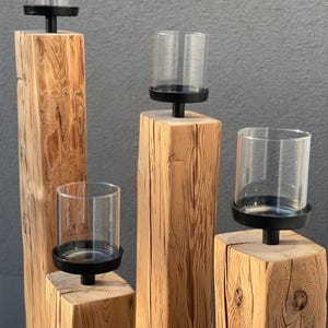 K&ouml;nnte beinhalten: Vier h&ouml;lzerne Kerzenhalter mit transparenten Glas-Windlichtern. Die Kerzenhalter sind aus Naturholz mit rauer Textur gefertigt. Jeder Halter hat einen schwarzen Metallsockel, um den Glasschirm zu halten. Der Hintergrund ist einfarbig grau.