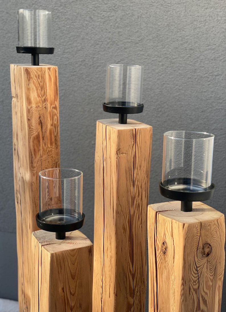 K&ouml;nnte beinhalten: Vier h&ouml;lzerne Kerzenhalter mit transparenten Glas-Windlichtern. Die Kerzenhalter sind aus Naturholz mit rauer Textur gefertigt. Jeder Halter hat einen schwarzen Metallsockel, um den Glasschirm zu halten. Der Hintergrund ist einfarbig grau.