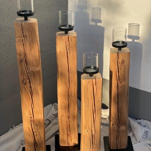 K&ouml;nnte beinhalten: Vier rustikale Kerzenhalter aus Holz. Jeder hat einen quadratischen, nat&uuml;rlichen Holzsockel mit einem schwarzen Metallsockel. Auf jedem Halter befindet sich ein klarer Glaszylinder, perfekt f&uuml;r Kerzen. Die Holzmaserung ist sichtbar.