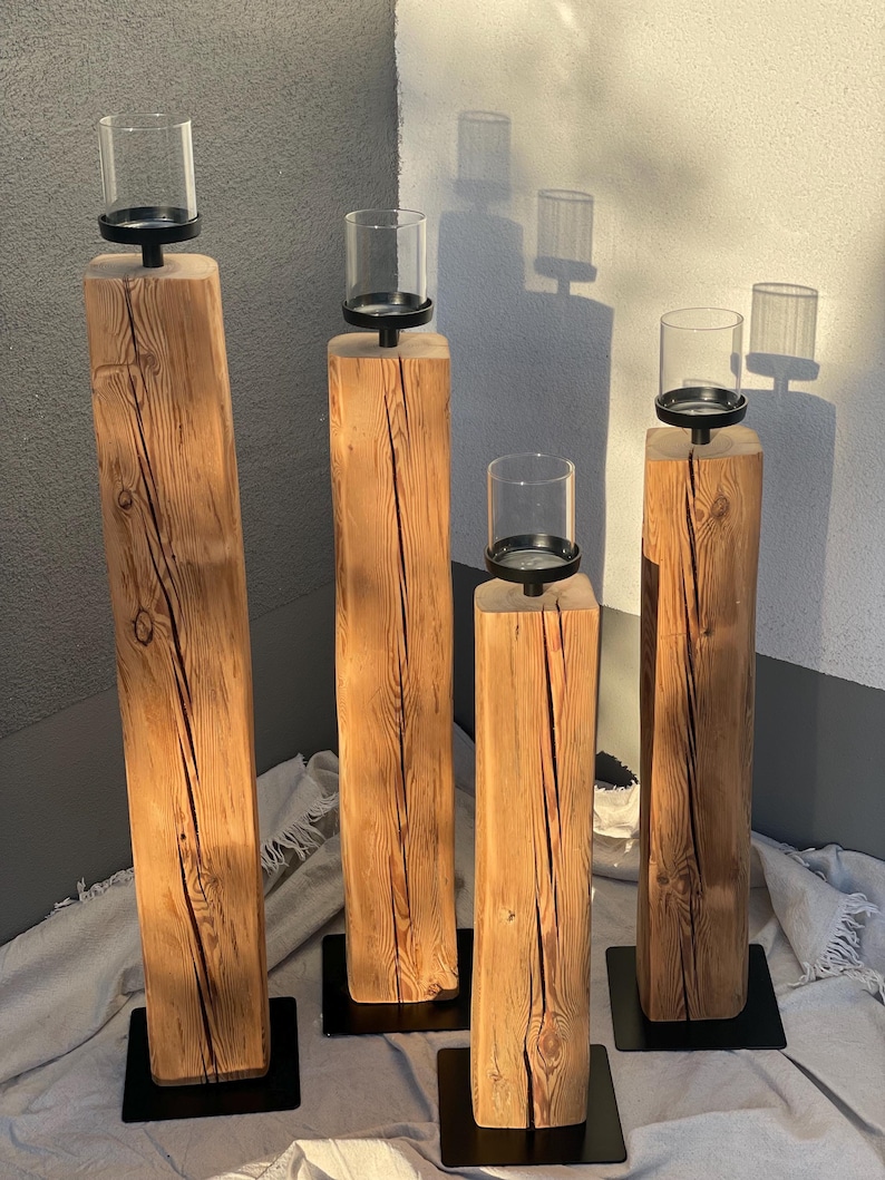 K&ouml;nnte beinhalten: Vier rustikale Kerzenhalter aus Holz. Jeder hat einen quadratischen, nat&uuml;rlichen Holzsockel mit einem schwarzen Metallsockel. Auf jedem Halter befindet sich ein klarer Glaszylinder, perfekt f&uuml;r Kerzen. Die Holzmaserung ist sichtbar.
