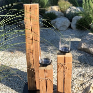 K&ouml;nnte beinhalten: Drei Kerzenhalter aus Holz in verschiedenen H&ouml;hen. Jeder Halter hat eine klare Glas-Teelichtschale auf einem schwarzen Metallsockel. Das Holz hat ein nat&uuml;rliches, verwittertes Aussehen mit sichtbarer Maserung und Knoten. Der kleinste Halter ist mit Bindfaden gebunden.