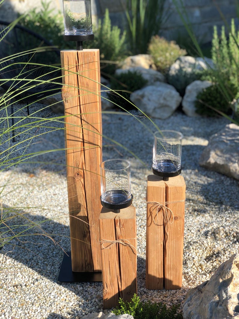 K&ouml;nnte beinhalten: Drei Kerzenhalter aus Holz in verschiedenen H&ouml;hen. Jeder Halter hat eine klare Glas-Teelichtschale auf einem schwarzen Metallsockel. Das Holz hat ein nat&uuml;rliches, verwittertes Aussehen mit sichtbarer Maserung und Knoten. Der kleinste Halter ist mit Bindfaden gebunden.