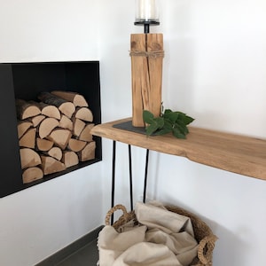 K&ouml;nnte beinhalten: Ein rustikaler Kerzenhalter aus Holz mit einem Glas-Windlichtaufsatz steht auf einem Holztisch mit schwarzen Haarnadelbeinen. Ein geflochtener Korb mit schwarzem Boden enth&auml;lt eine cremefarbene Decke. Brennholz ist in einer schwarzen, eingelassenen Wandnische gestapelt.