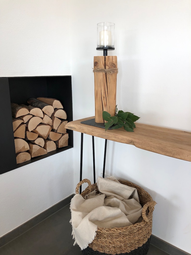 K&ouml;nnte beinhalten: Ein rustikaler Kerzenhalter aus Holz mit einem Glas-Windlichtaufsatz steht auf einem Holztisch mit schwarzen Haarnadelbeinen. Ein geflochtener Korb mit schwarzem Boden enth&auml;lt eine cremefarbene Decke. Brennholz ist in einer schwarzen, eingelassenen Wandnische gestapelt.