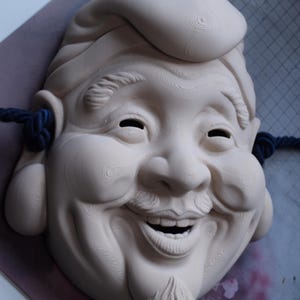 May include: A carved, off-white wooden mask depicting a smiling, elderly man with a mustache and goatee. The mask has a prominent forehead, closed eyes, and a jovial expression. Dark blue rope is attached to the sides.