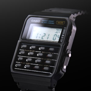 May include: A black Casio CA53W-1 calculator watch with a digital display showing the time 1:21:04. The watch has a black band and a black case. The watch has a variety of buttons for setting the time, alarm, and stopwatch.